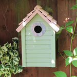 New England Nest Box - The Irish Gardener Store New England Nest Box, green wooden birdhouse with horizontal slats, natural wood-colored pitched roof, circular black metal entrance hole, mounted on a wooden fence, surrounded by lush green foliage, durable and stylish bird nesting box for garden birds