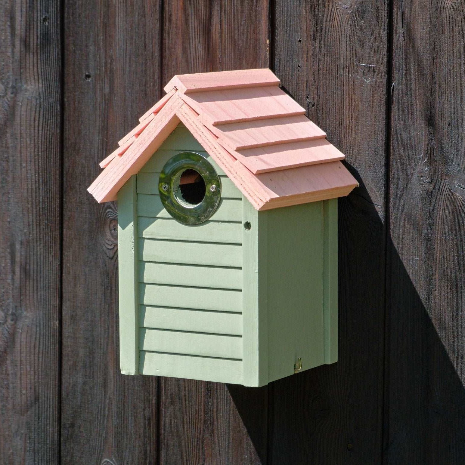 New England Nest Box - The Irish Gardener Store New England Nest Box, green wooden birdhouse with horizontal slats, natural wood-colored pitched roof, circular black metal entrance hole, mounted on a wooden fence, durable and stylish bird nesting box for garden birds