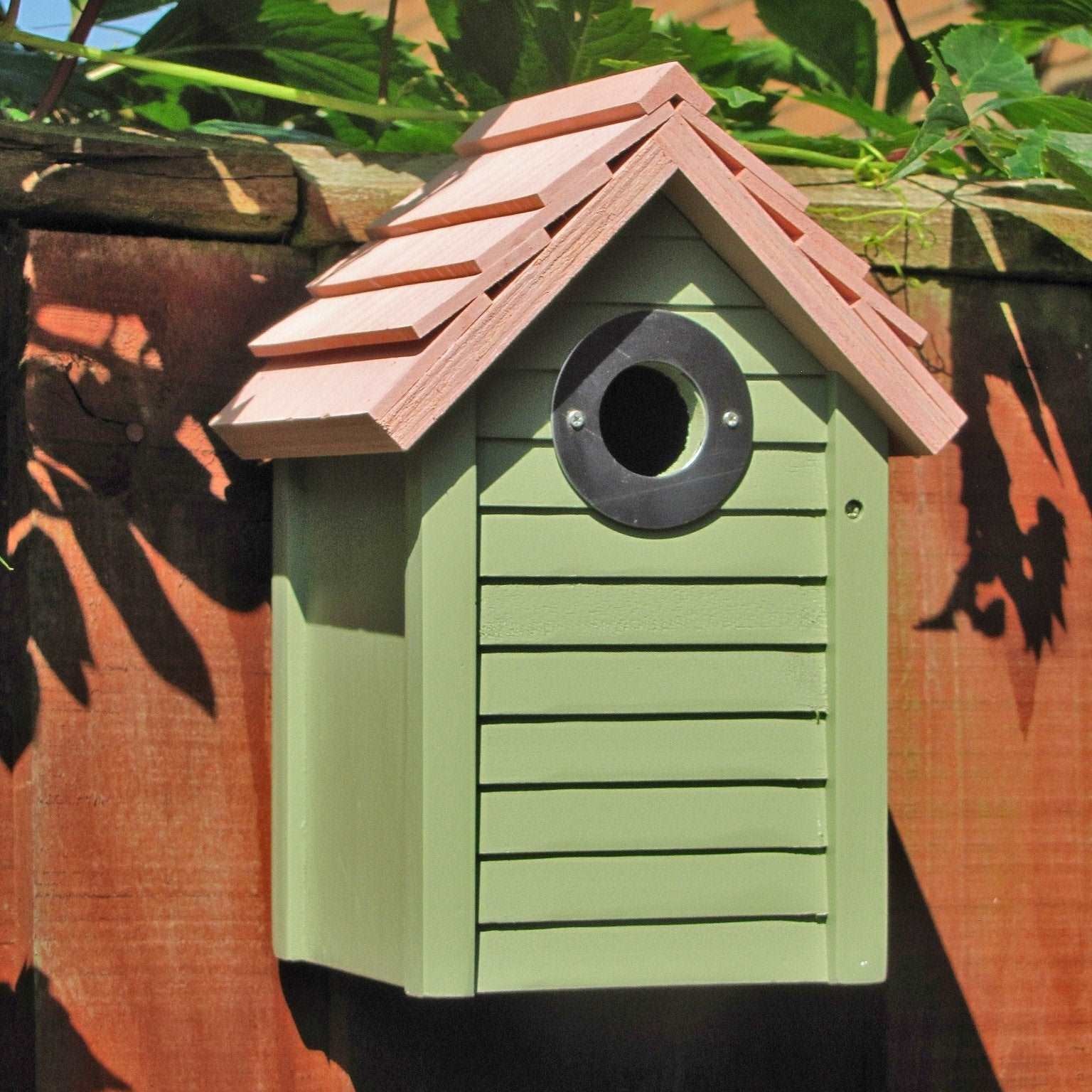 New England Nest Box - The Irish Gardener Store New England Nest Box, green wooden birdhouse with horizontal slats, natural wood-colored pitched roof, circular black metal entrance hole, mounted on a wooden fence, surrounded by lush green foliage, durable and stylish bird nesting box for garden birds