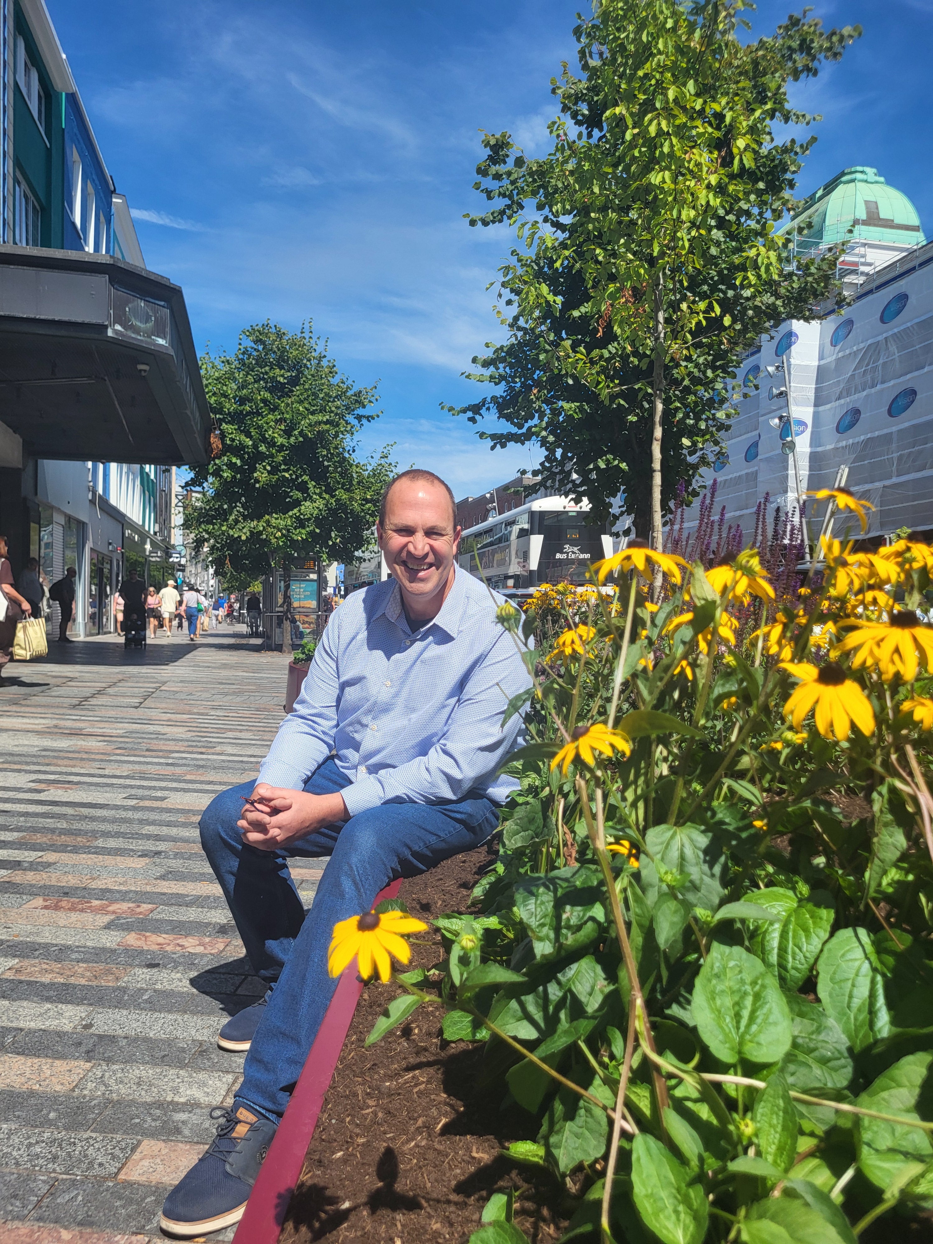 Peter Dowdall, Irish horticulturist and broadcaster, sitting beside yellow rudbeckia planting on St Patrick's Street, Cork, an urban greening pilot with trees and seating