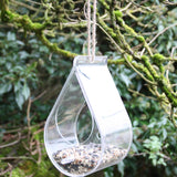 Hanging dewdrop-shaped bird feeder, made of clear acrylic, filled with birdseed, suspended from a moss-covered tree branch using a natural rope, surrounded by lush greenery and woodland environment, eco-friendly and decorative feeder design