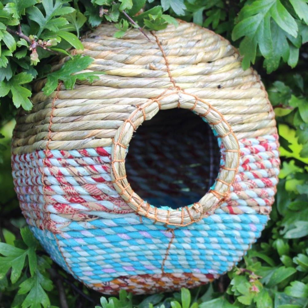 A spherical Anita Artisan Bird Nester, woven from natural materials, featuring colorful blue, red, and beige patterns, a round central entrance hole, securely attached to green foliage, designed as a cozy and decorative shelter for small garden birds.