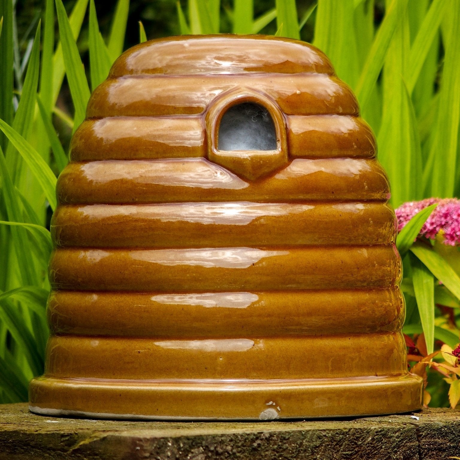 Ceramic Bumblebee Nester, beehive-shaped design, glossy brown ceramic finish, small entrance hole, placed on a garden surface, surrounded by green foliage and colorful flowers, eco-friendly pollinator habitat






