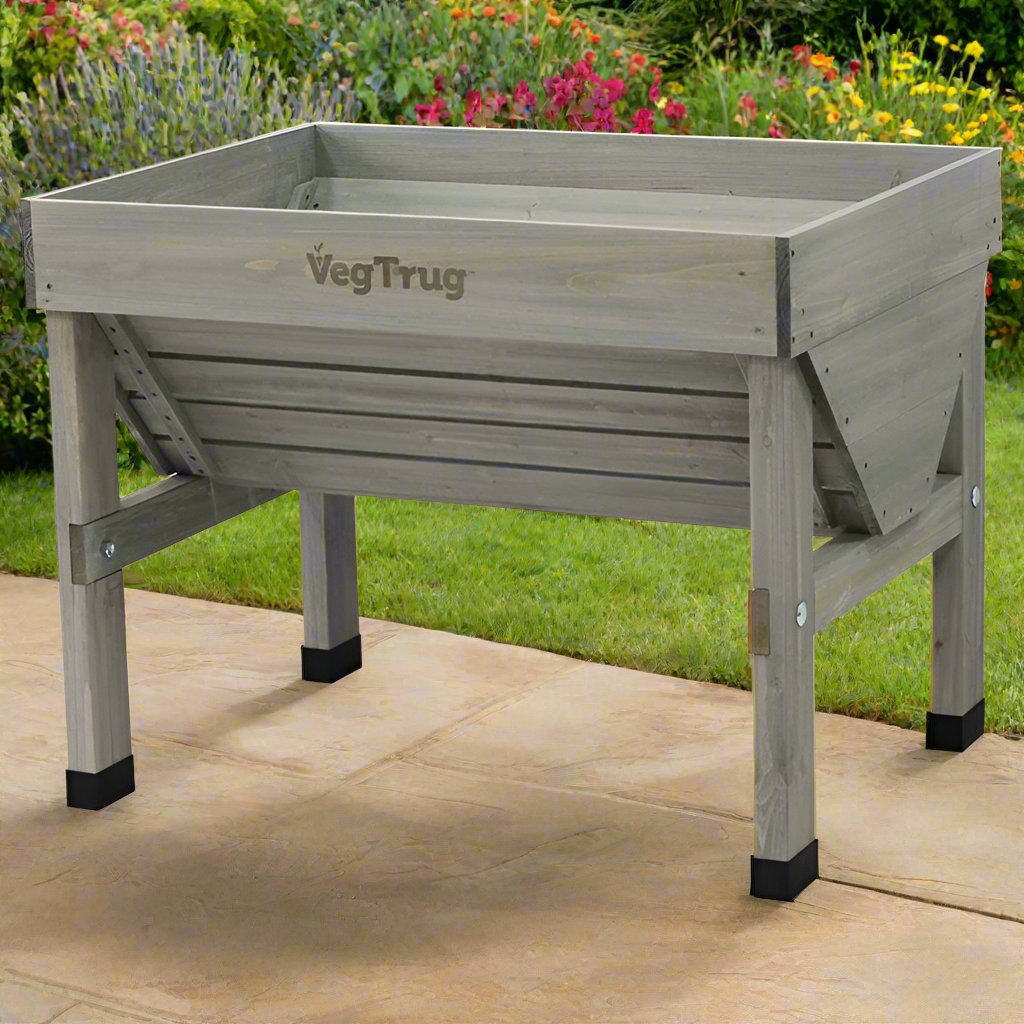 A grey wooden VegTrug raised garden bed with a slatted design, elevated on sturdy legs with black protective caps, positioned on a paved patio, surrounded by a lush garden with colorful flowers and greenery in the background, ideal for comfortable and convenient gardening.